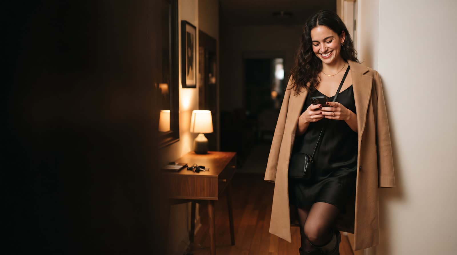 A woman in her hallway, coat on and ready to go out, smiling at a message on her phone.