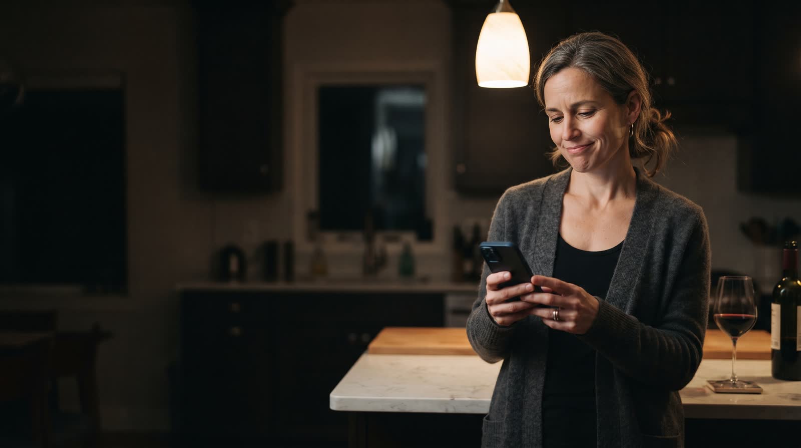 A woman at her kitchen counter in the evening, looking at her phone with an unsure half-smile.