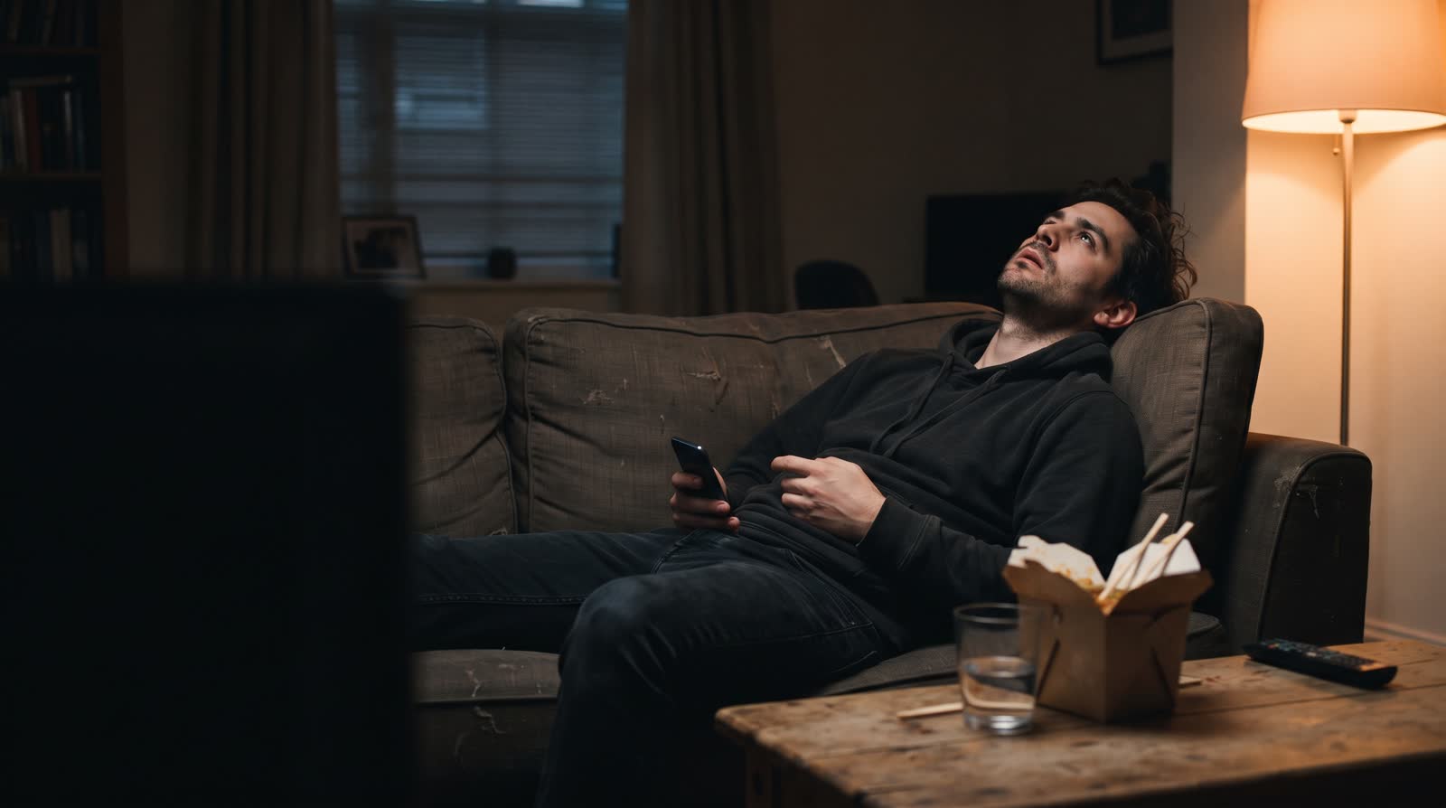 A man slumped on a sofa at night staring at the ceiling, phone in his hand.